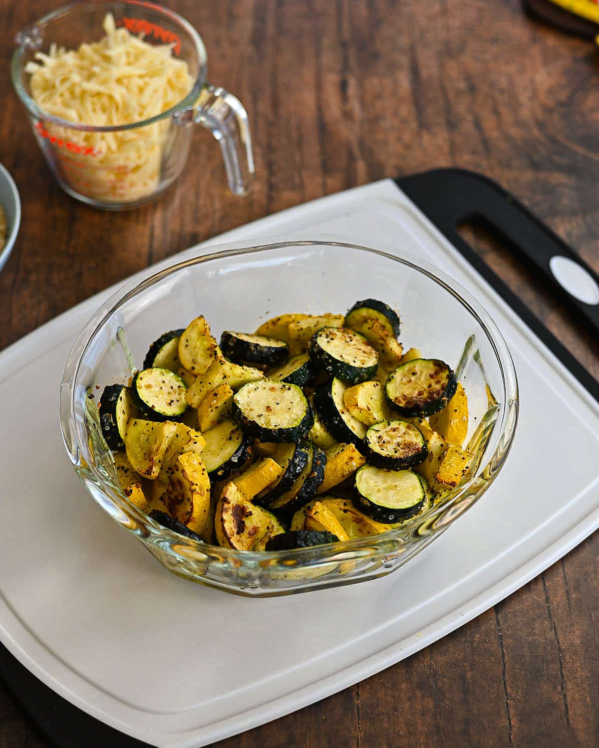 Slightly baked zucchini and yellow squash in a clear casserole dish sitting on a white cutting board.