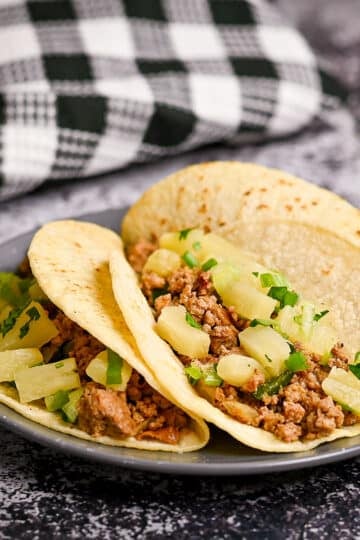 Three tortillas filled with ground meat and pineapple on a gray plate.