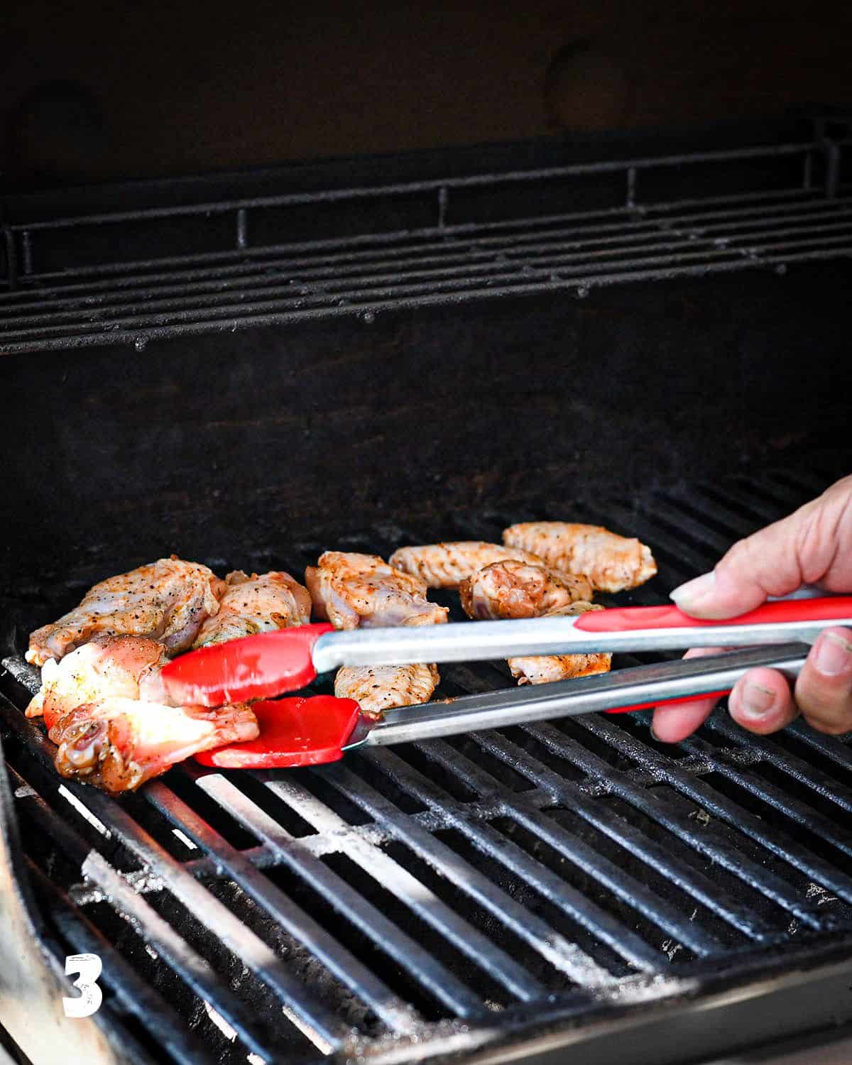 Using red tongs to place chicken wings on the grill. 