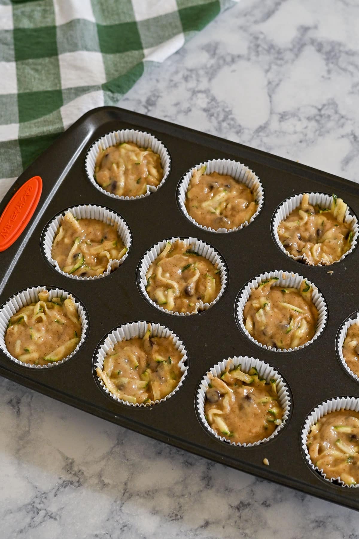 Muffin pan with paper liners filled with unbaked zucchini bread muffins batter on a marble counter, green checkered towel nearby.