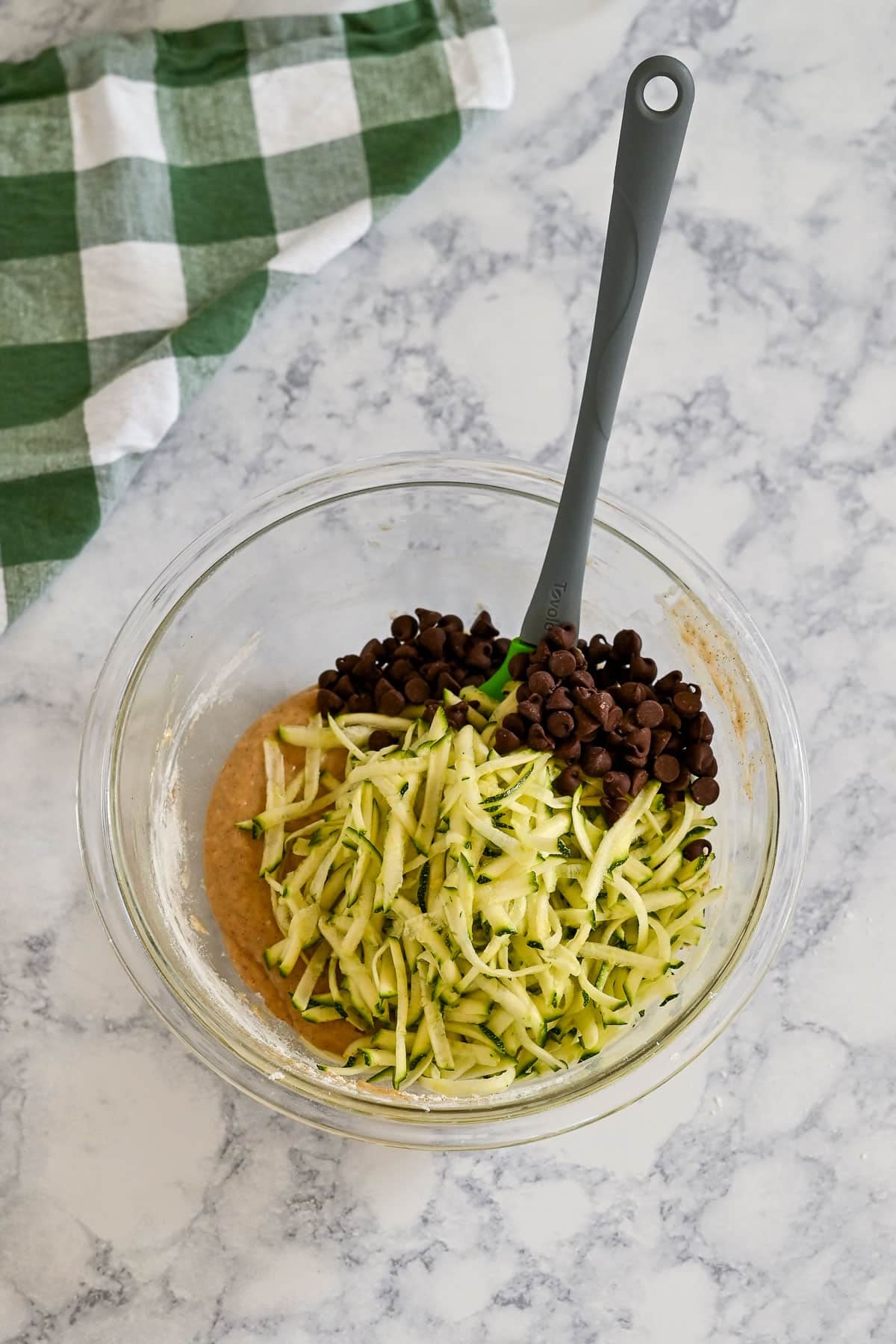 Clear glass bowl with shredded zucchini and chocolate chips on batter, perfect for making zucchini bread muffins with applesauce, on a marble surface with a green checkered towel.