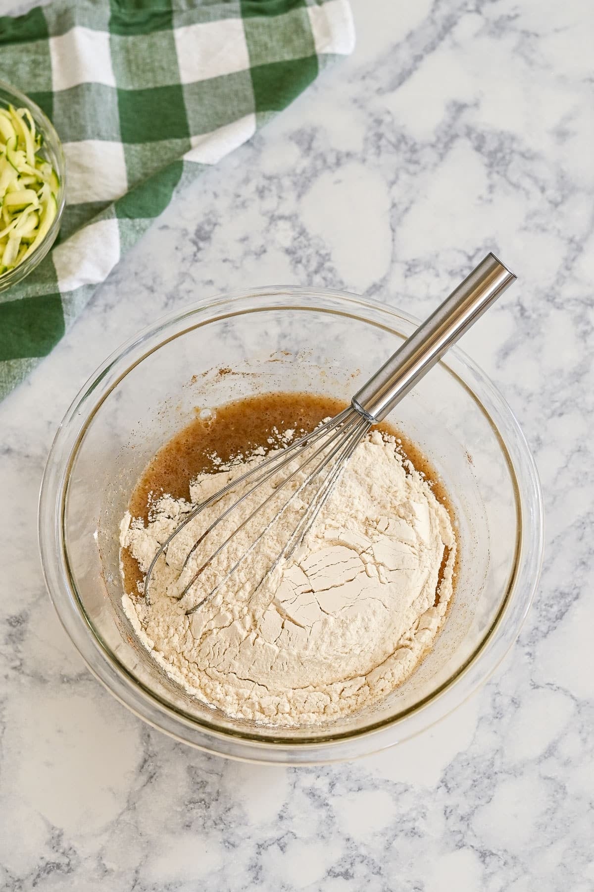 A glass bowl with flour and batter for zucchini bread muffins being mixed with a whisk on a marble countertop near a green checkered towel.
