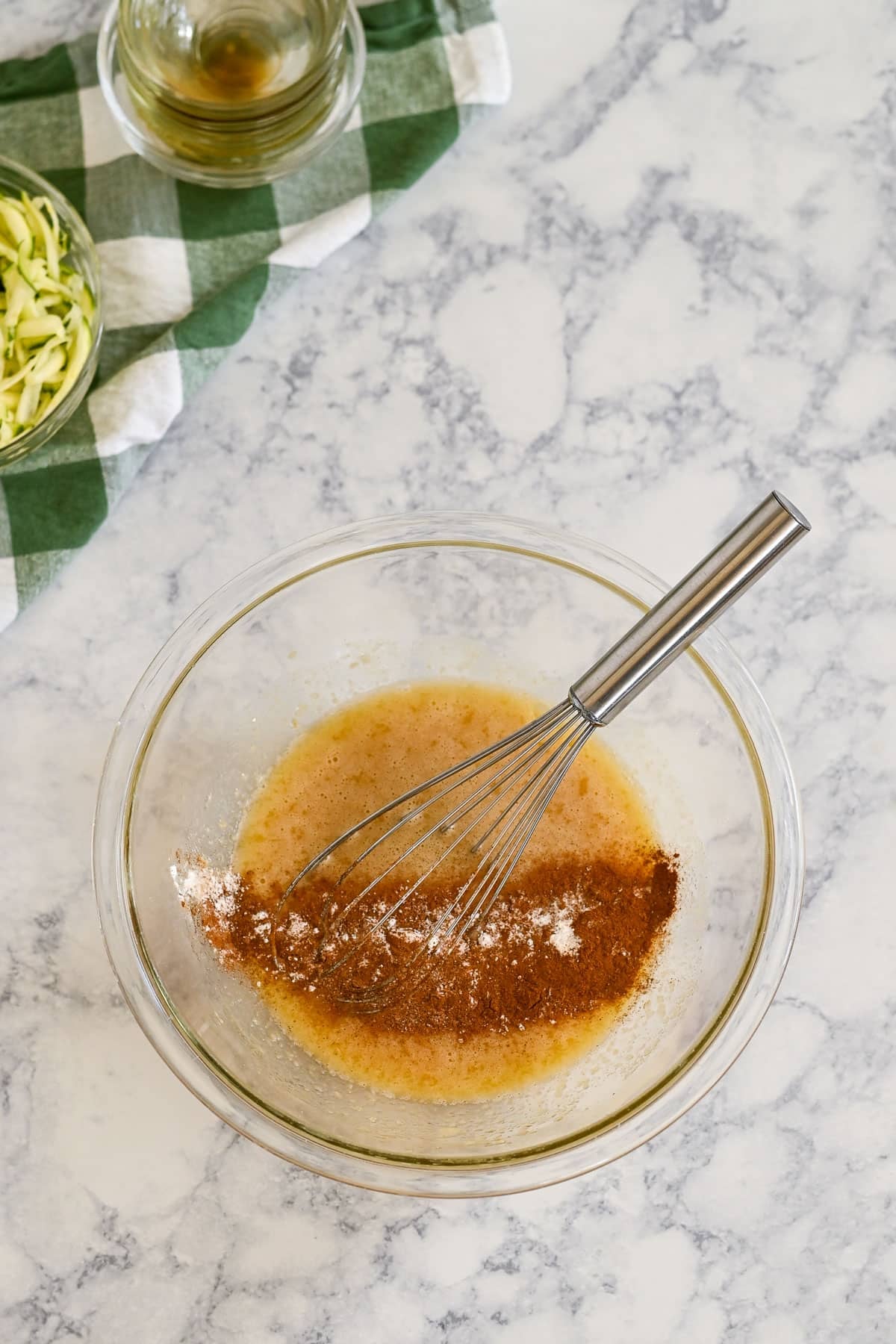 A glass bowl with whisked ingredients and cinnamon, perfect for making zucchini bread muffins with applesauce, sits on a marble counter with a whisk inside.