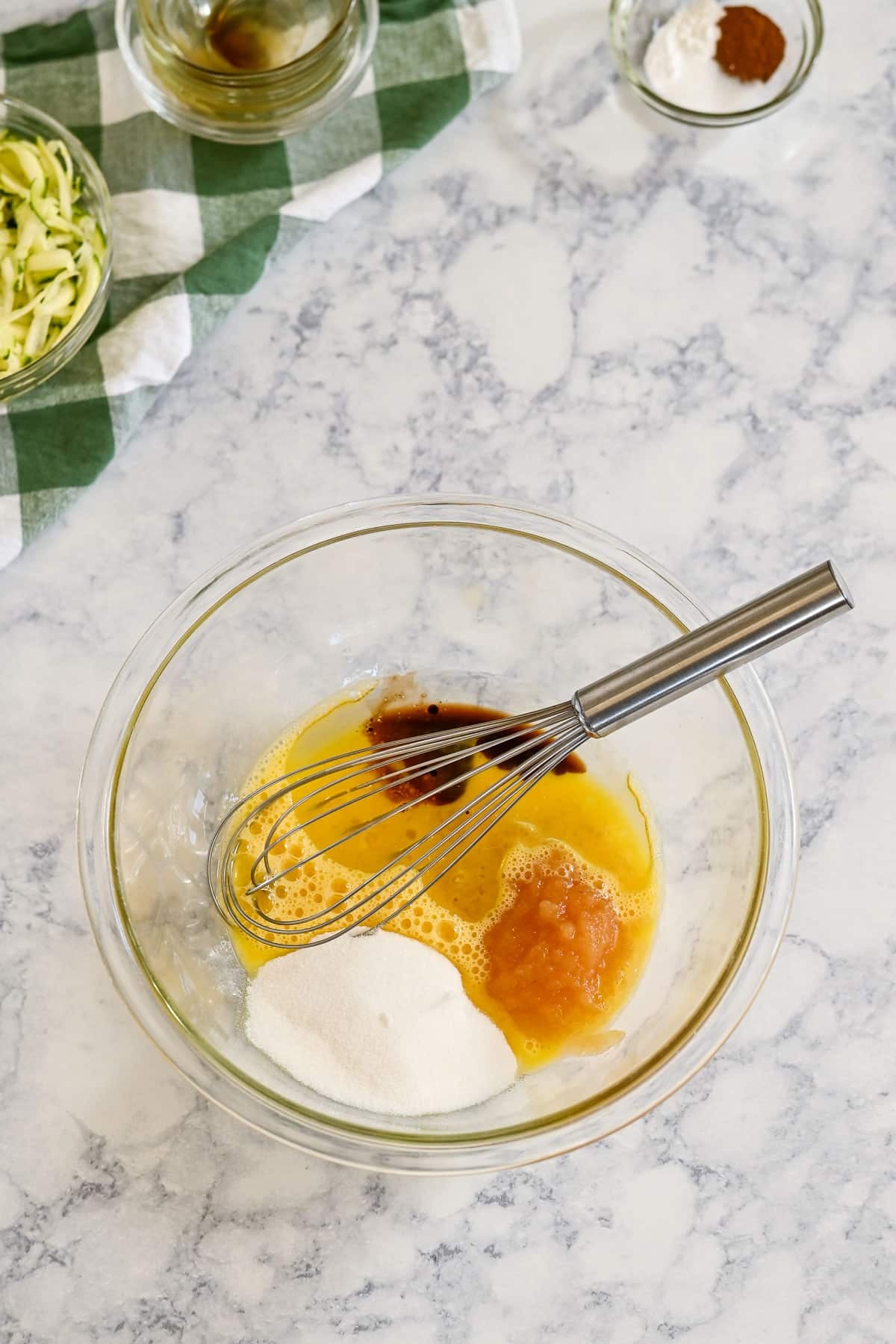 A glass bowl with eggs, sugar, applesauce, and vanilla being whisked on a marble counter for delicious zucchini bread muffins with applesauce.