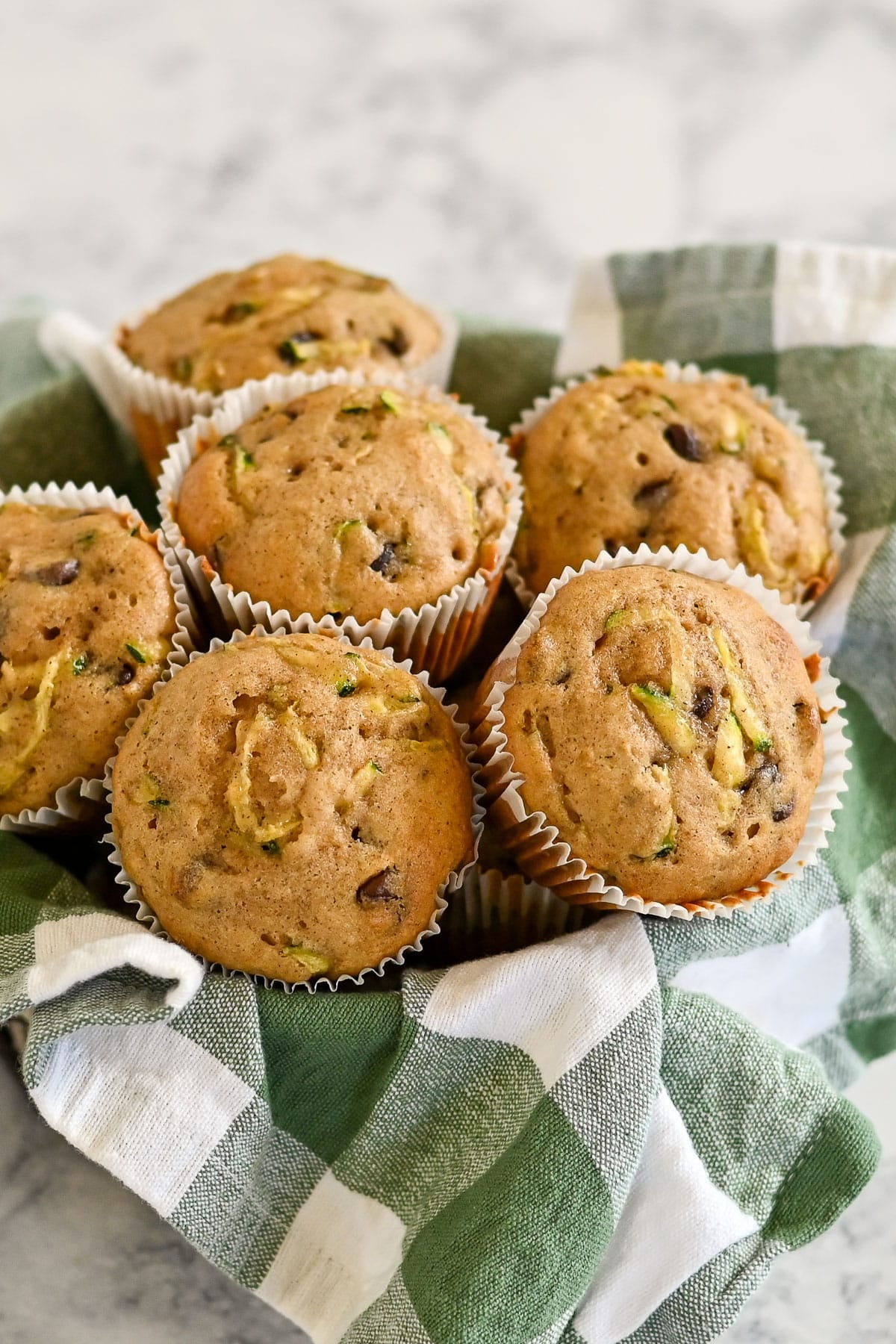 A basket of Zucchini Bread Muffins with applesauce and chocolate chips in white paper liners rests on a green and white checkered cloth.