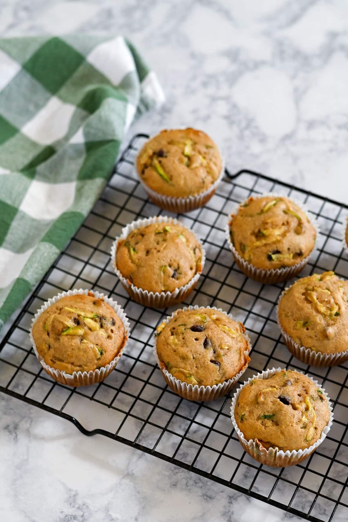 Seven Zucchini Bread Muffins with applesauce rest on a cooling rack beside a green and white checkered cloth on a marble surface.