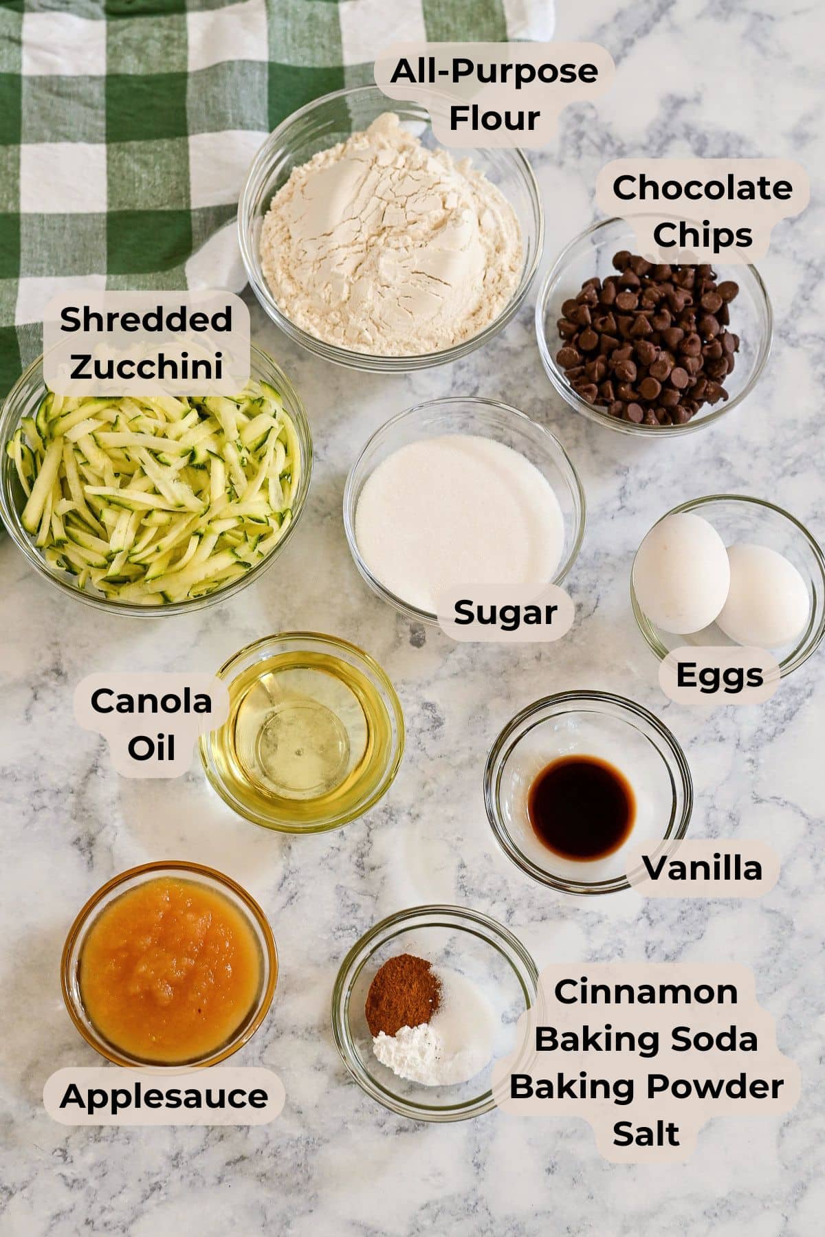 Ten labeled bowls with ingredients for baking zucchini bread muffins: flour, chocolate chips, zucchini, sugar, eggs, oil, vanilla, applesauce, and spices-everything you need for delicious zucchini bread muffins with applesauce.