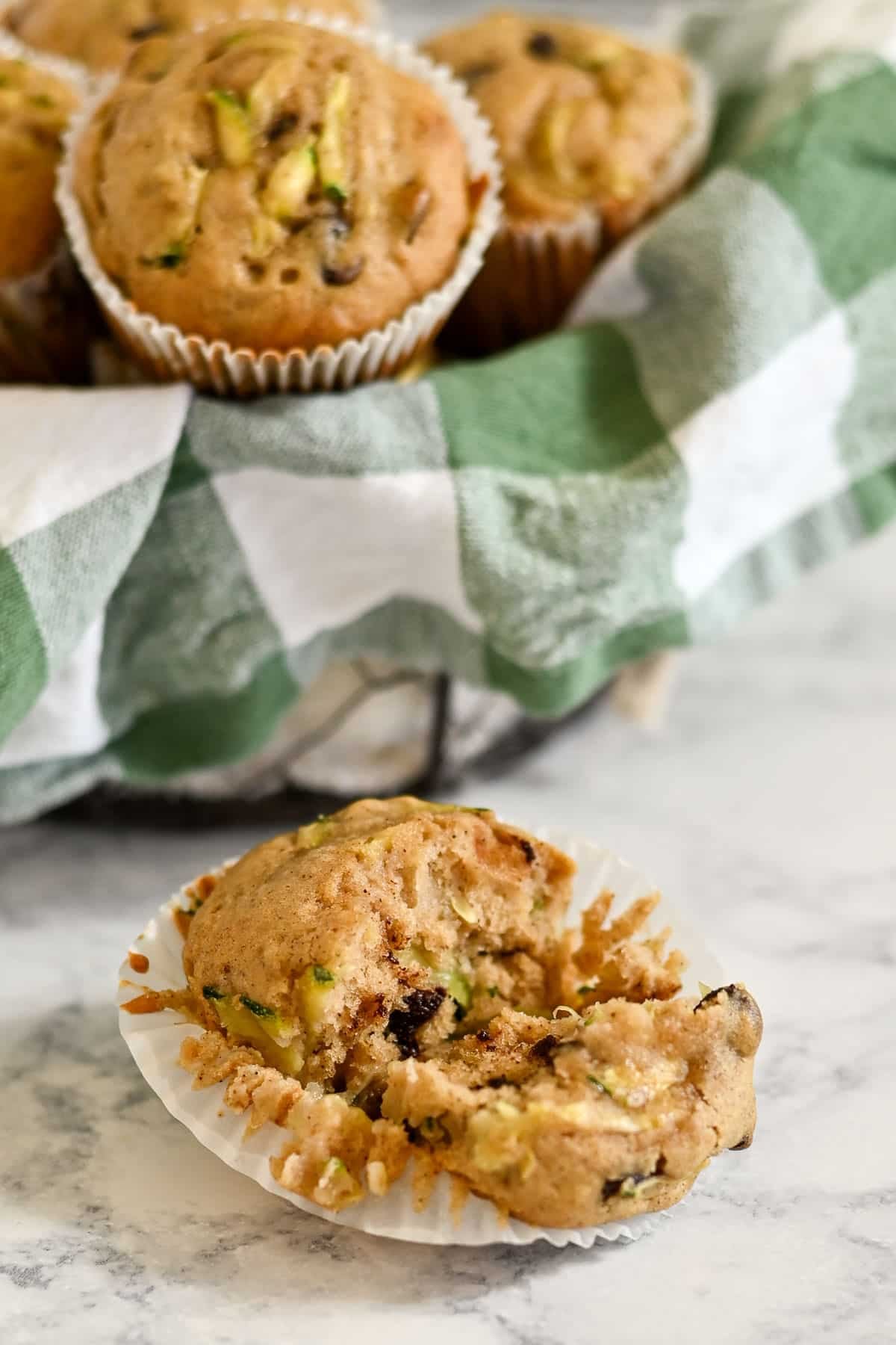 A bitten zucchini bread muffin in a wrapper, with more zucchini bread muffins with applesauce arranged in a basket lined with a green and white cloth.