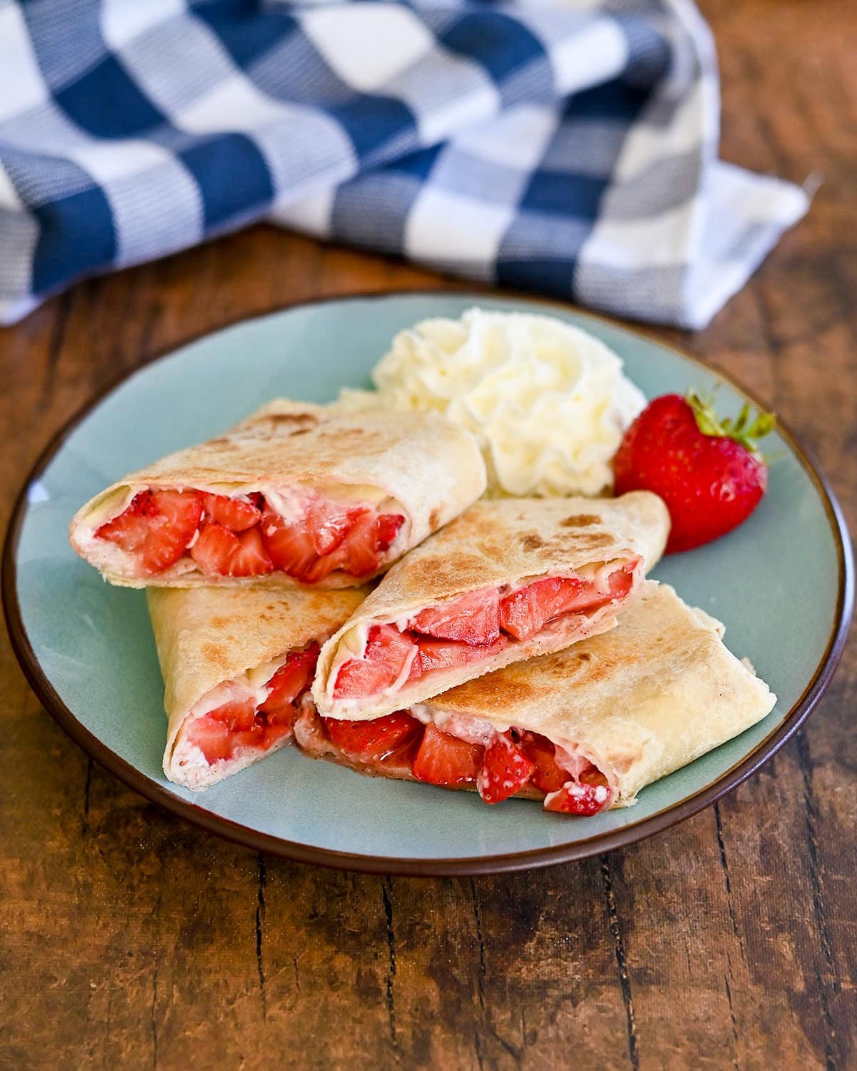 Finished strawberry burritos on a plate with whipped cream next to a checkered towel.