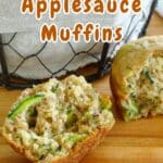Zucchini bread muffins with applesauce rest on a wooden surface, one revealing its moist interior dotted with tender zucchini pieces.