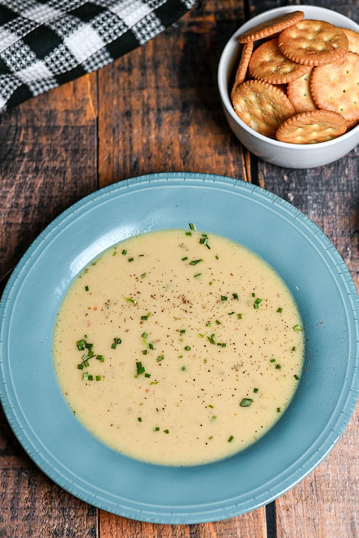 Creamy soup with herbs in a blue bowl, next to a bowl of round crackers on a wooden table.