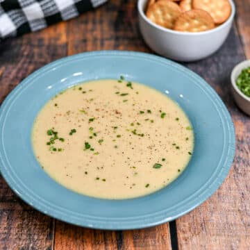 Creamy homemade cream of onion soup garnished with chives in a blue bowl, served alongside crackers and fresh herbs in bowls nearby on a wooden table-an inviting presentation that showcases the essential ingredients for cream of onion soup.