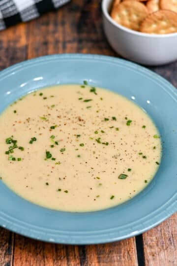 Creamy homemade cream of onion soup garnished with chives in a blue bowl, served alongside crackers and fresh herbs in bowls nearby on a wooden table-an inviting presentation that showcases the essential ingredients for cream of onion soup.