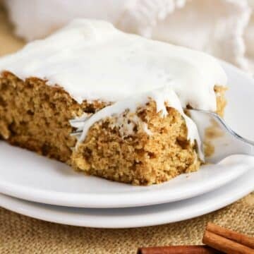 A fork cuts into a thick slice of old-fashioned applesauce cake, generously frosted and served on a white plate, with cinnamon sticks nearby.