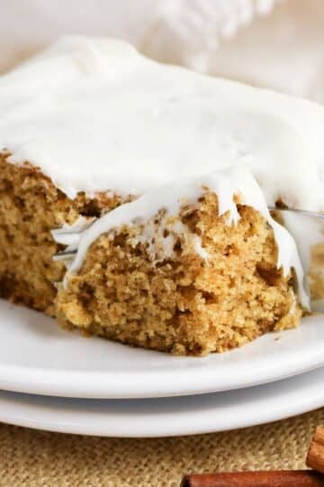A fork cuts into a thick slice of old-fashioned applesauce cake, generously frosted and served on a white plate, with cinnamon sticks nearby.