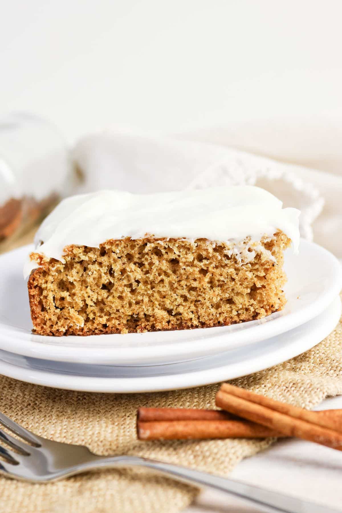 A slice of spiced Old-fashioned applesauce cake with white frosting sits on a white plate, accompanied by cinnamon sticks and a fork nearby.