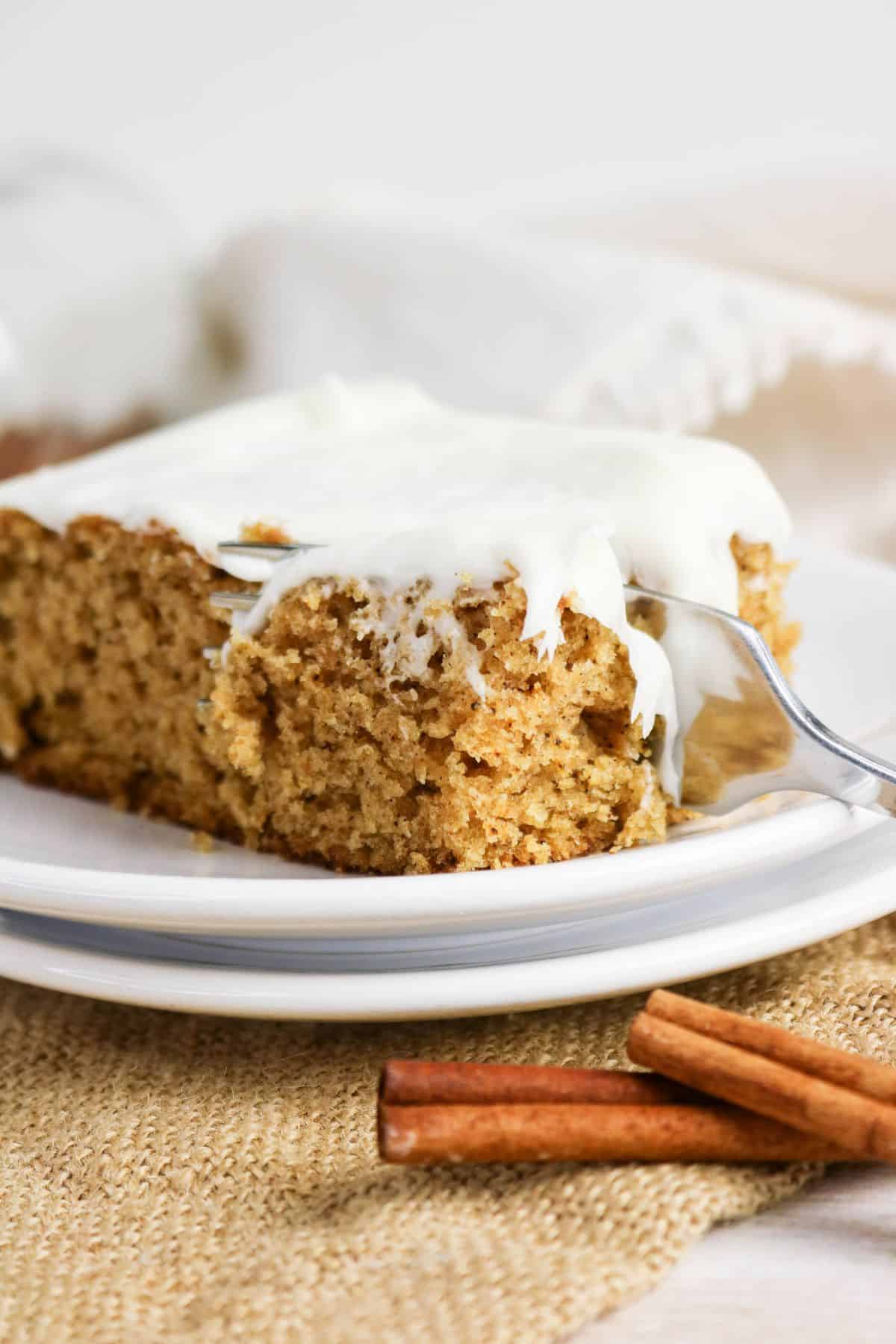 A slice of frosted, old-fashioned applesauce cake rests on a plate with a fork, next to cinnamon sticks on a woven mat.