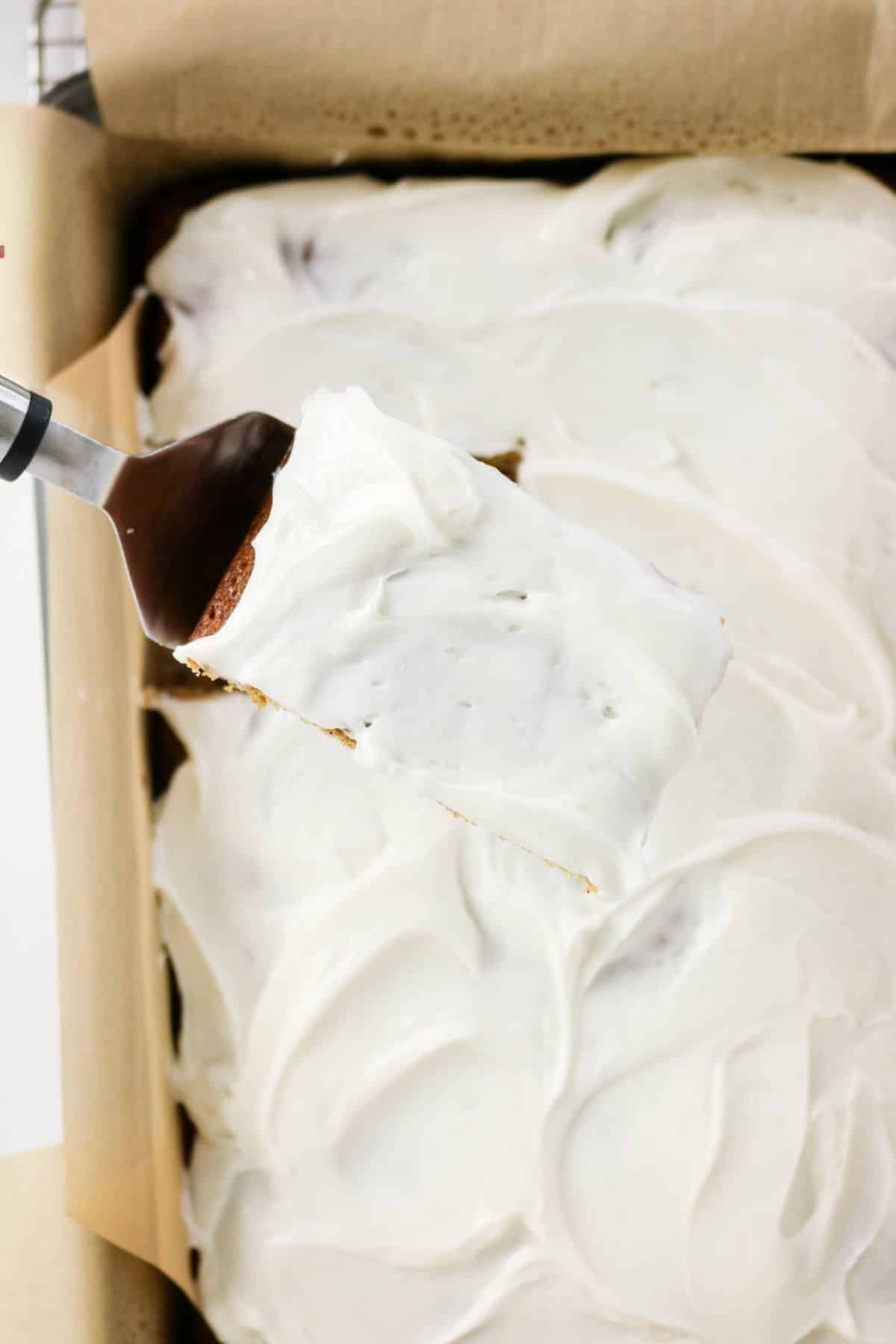 A spatula lifts a frosted slice of old-fashioned applesauce cake from a baking pan lined with parchment paper.