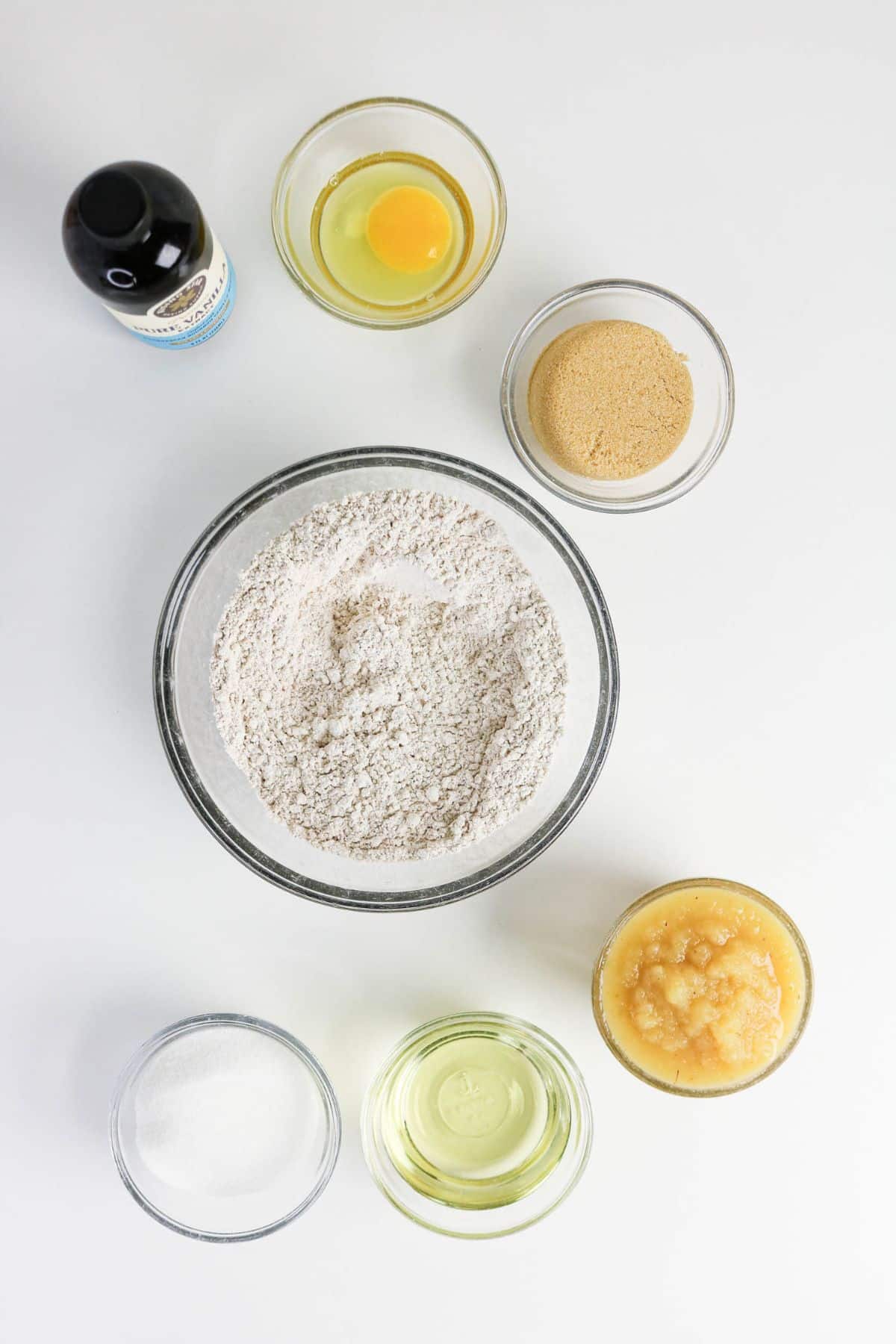 Seven baking ingredients in bowls-including flour, egg, sugars, applesauce, oil, and vanilla-are beautifully arranged on a white surface.