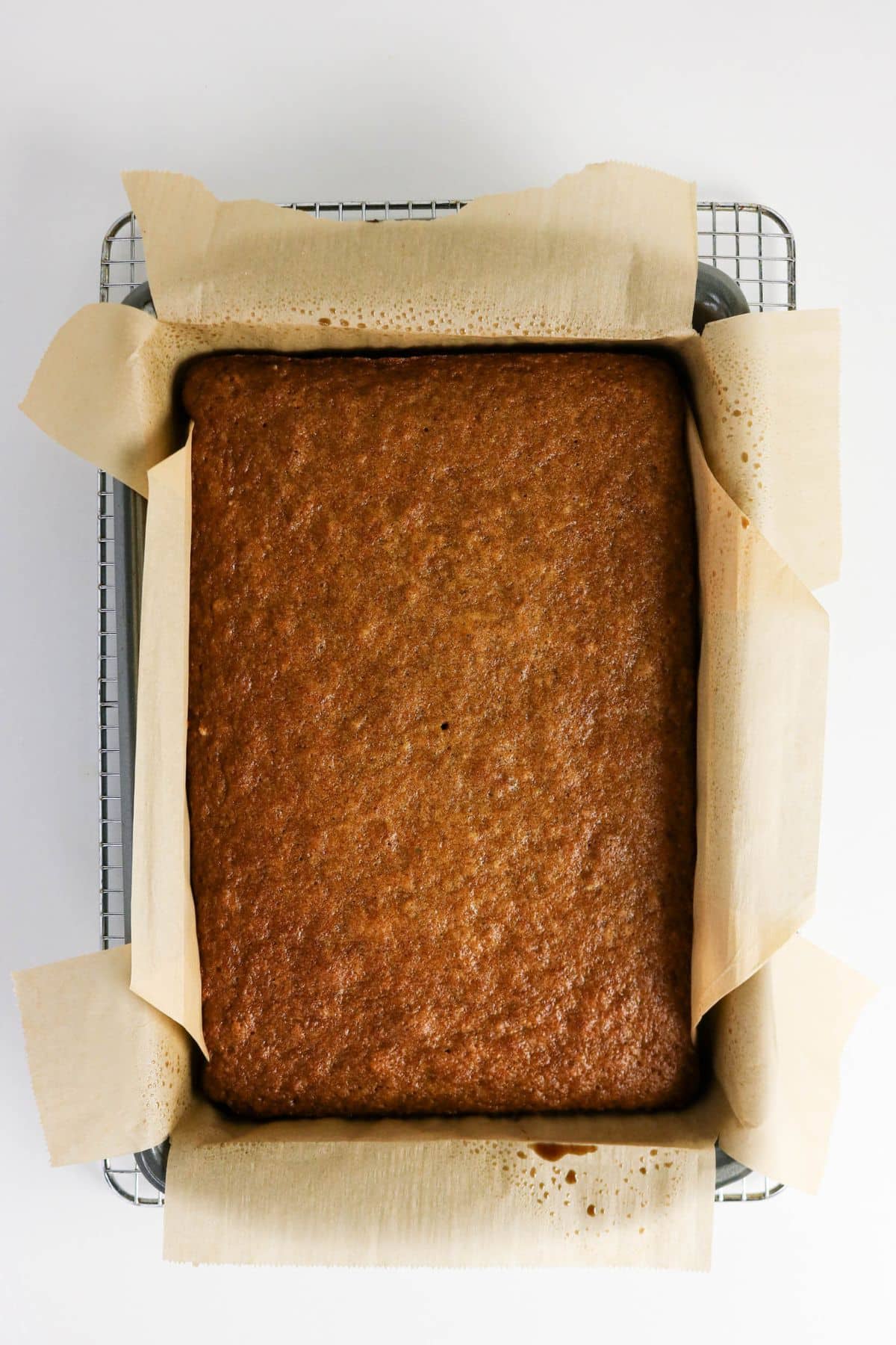 A rectangular Old-fashioned applesauce cake rests in a pan lined with parchment paper on a cooling rack.