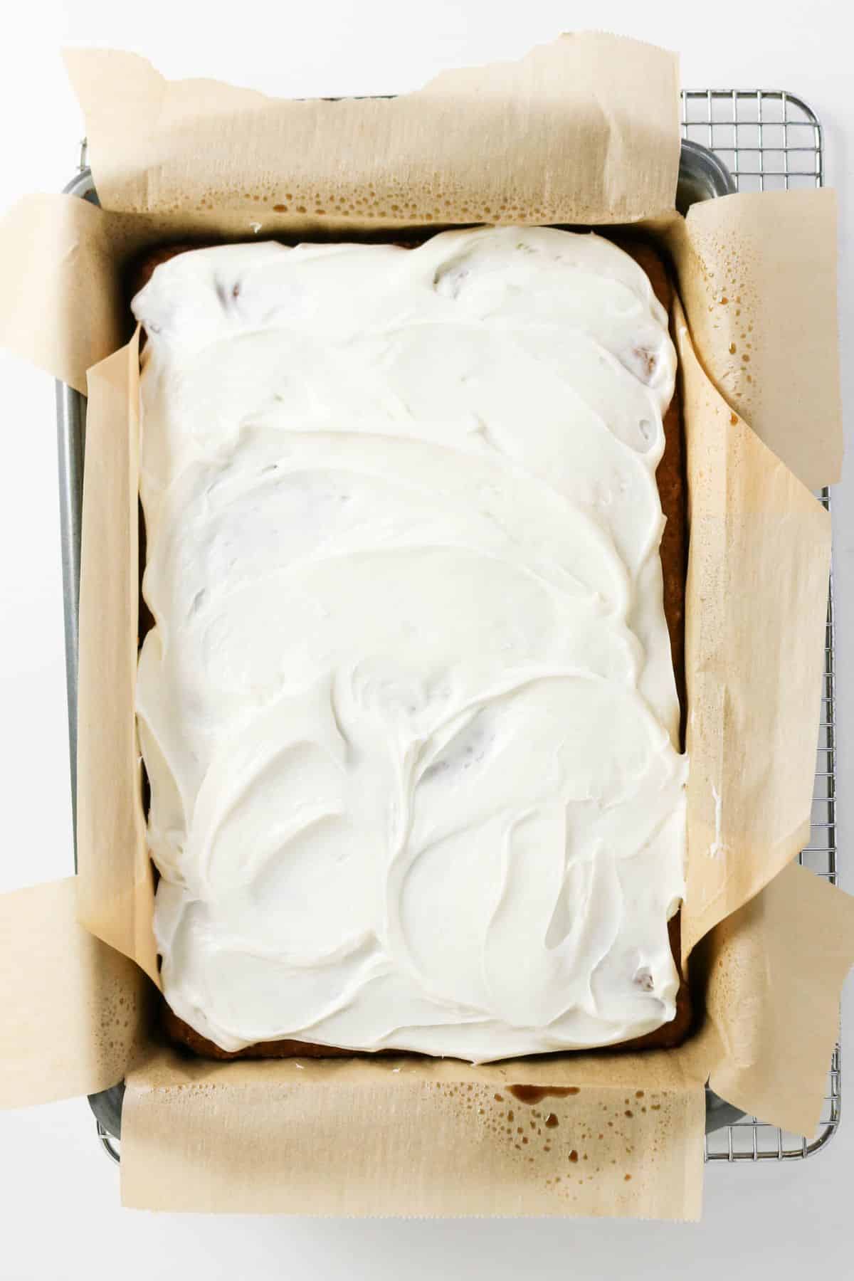 A rectangular Old-fashioned applesauce cake covered with white frosting sits in a parchment-lined baking pan on a cooling rack.