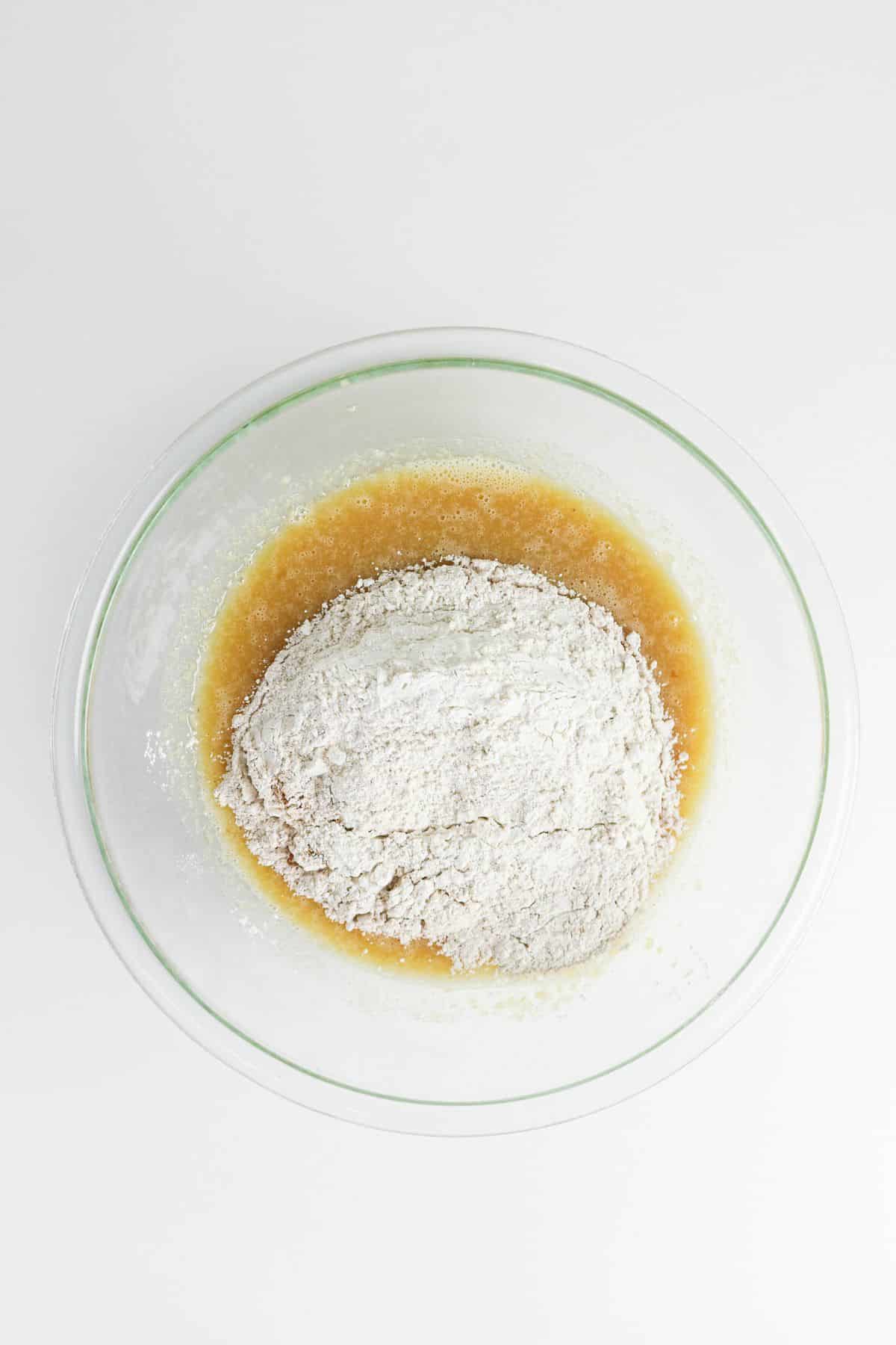Clear glass bowl with flour and wet ingredients being mixed, viewed from above on a white background.