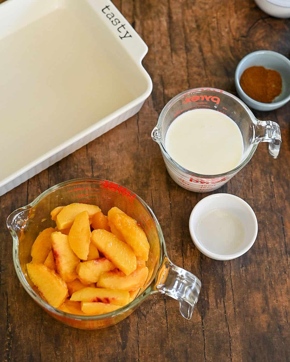 Sliced peaches, milk, and cinnamon are arranged in a small dish on a wooden table next to a white baking dish, ready to be transformed into a delicious Fresh Peach Cobbler.