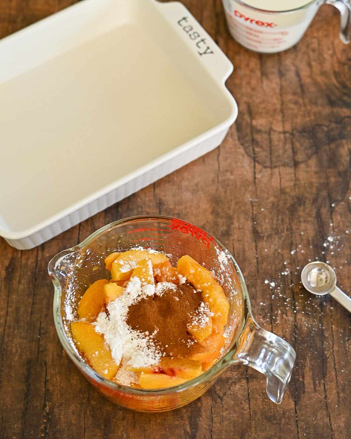 A glass measuring cup with sliced peaches, cinnamon, and cornstarch sits near a baking dish on a wooden table-perfect ingredients for making Fresh Peach Cobbler.