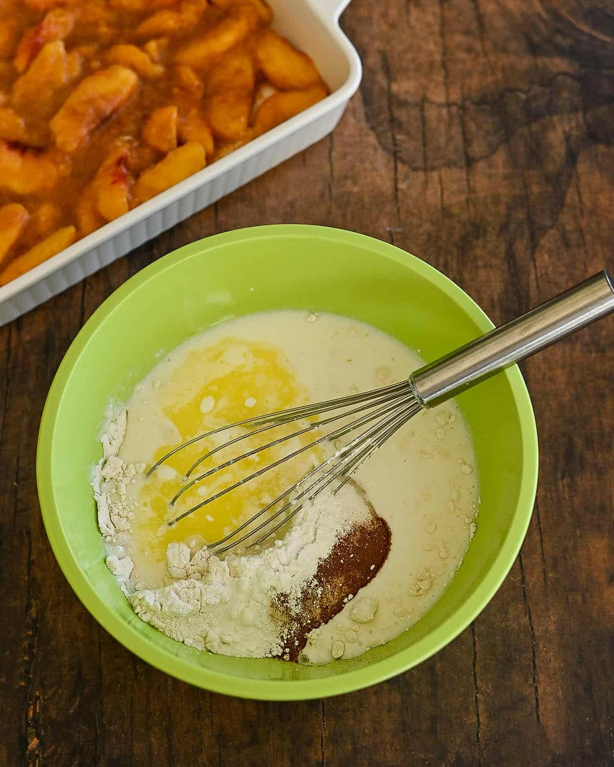 Green bowl with baking ingredients and a whisk, next to a dish of sliced peaches on a wooden surface-perfect for preparing a Fresh Peach Cobbler.