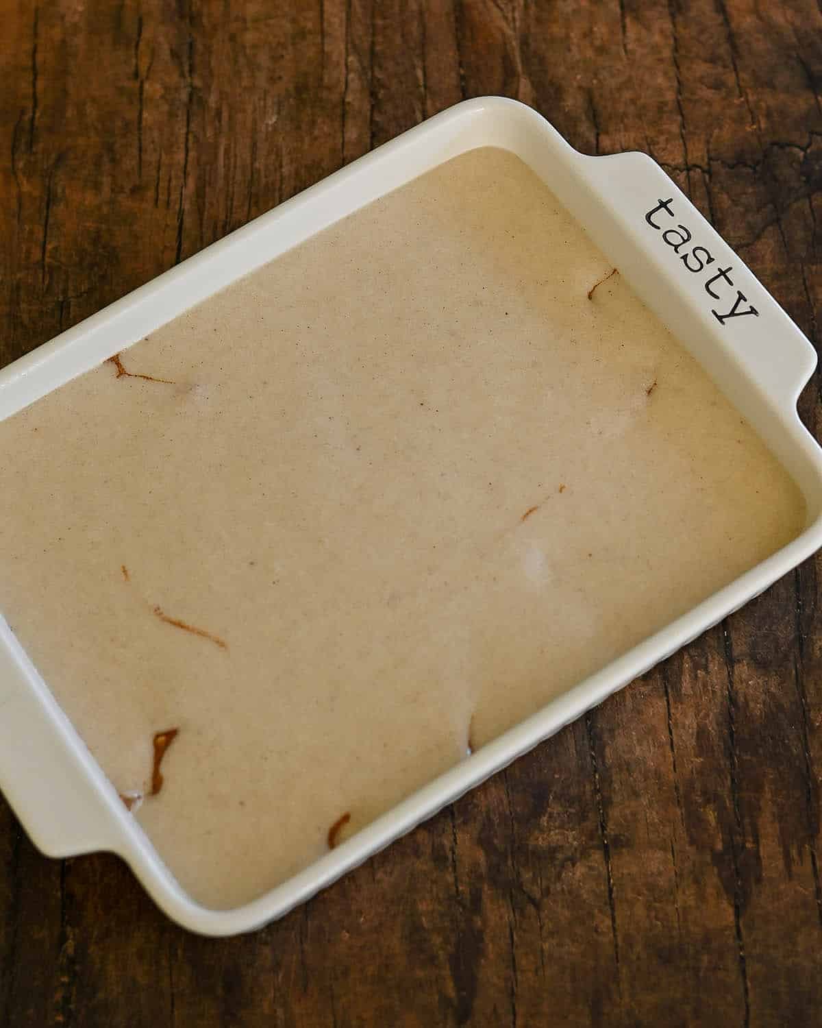Ceramic baking dish labeled "tasty" filled with light brown Fresh Peach Cobbler batter, resting on a wooden surface.