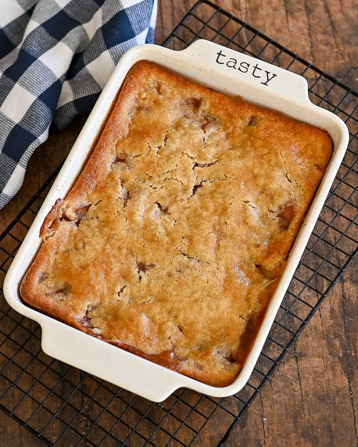 Golden brown Fresh Peach Cobbler in a white baking dish labeled "tasty," rests on a cooling rack with a plaid cloth nearby.