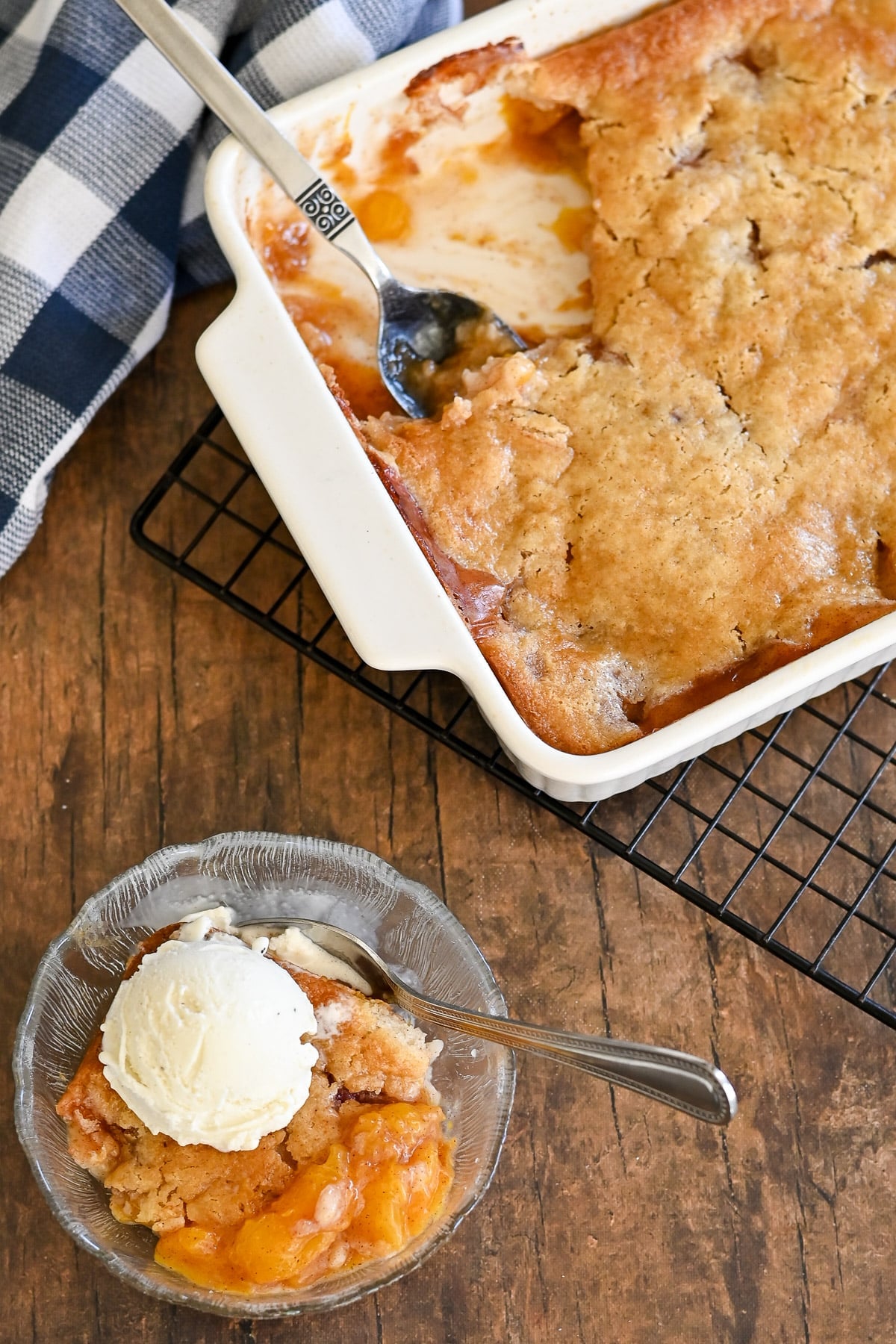 A bowl of fresh peach cobbler with vanilla ice cream sits beside a baking dish of cobbler on a cooling rack.