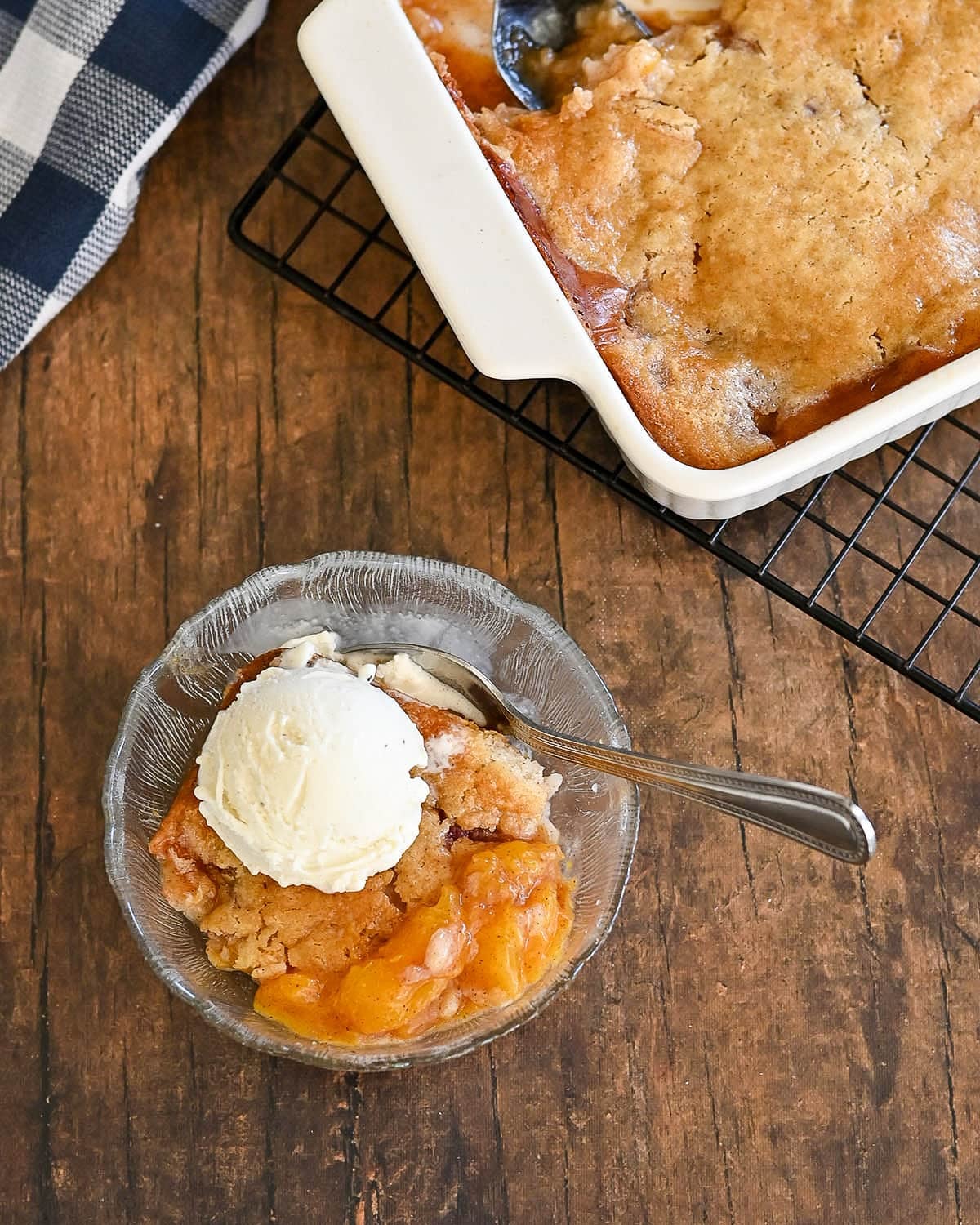 A bowl of Fresh Peach Cobbler with a scoop of vanilla ice cream sits next to a baking dish of cobbler on a wooden table.