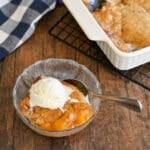 A glass bowl with Fresh Peach Cobbler and a scoop of vanilla ice cream, next to a baking dish on a wooden table.