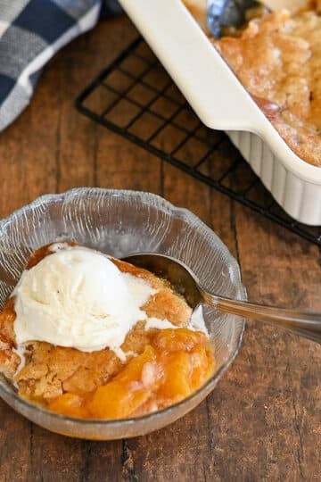 A glass bowl with Fresh Peach Cobbler and a scoop of vanilla ice cream, next to a baking dish on a wooden table.