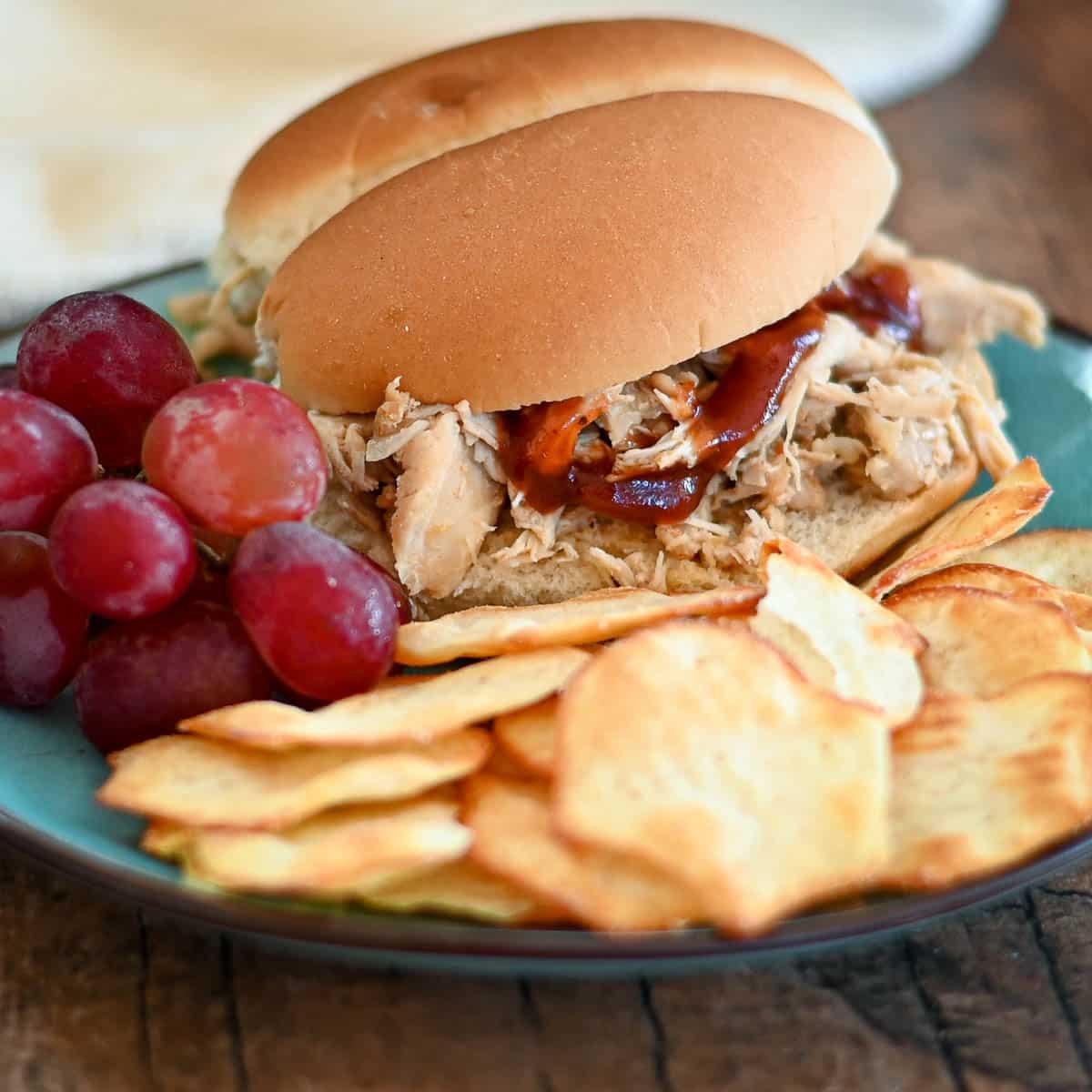 A shredded chicken sandwich next to chips and grapes on a plate.