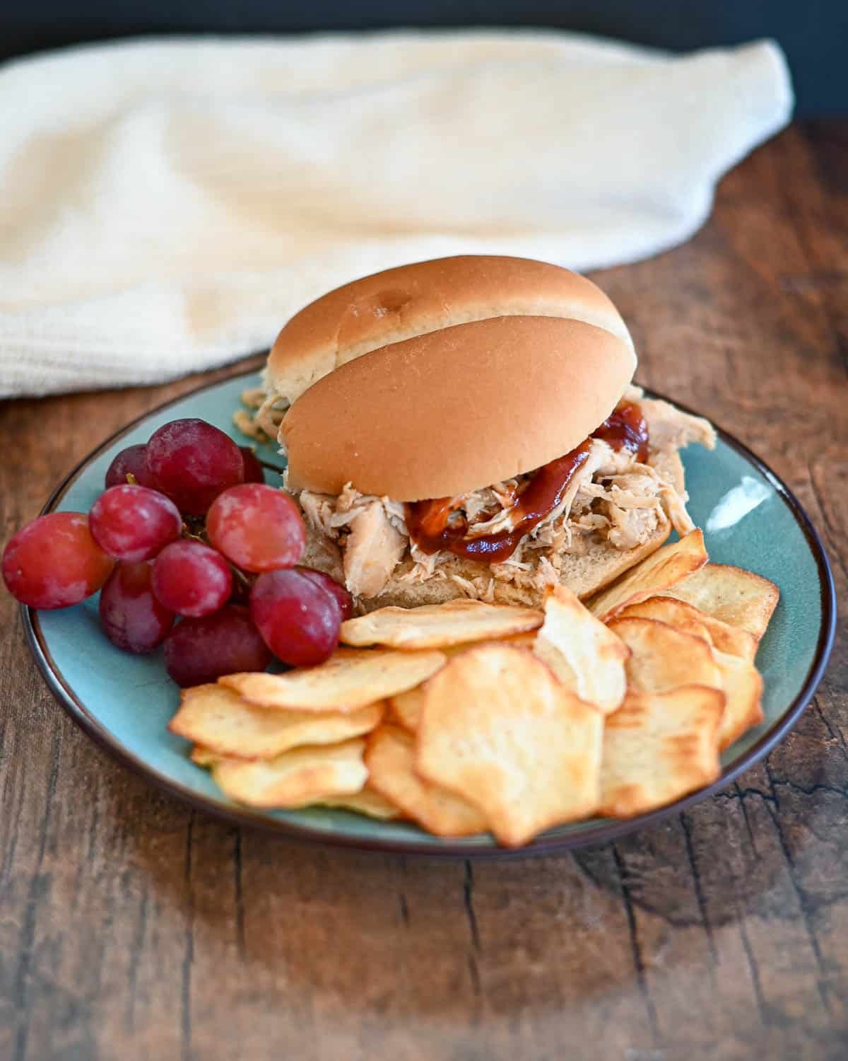 A shredded chicken sandwich next to chips and grapes on a plate.