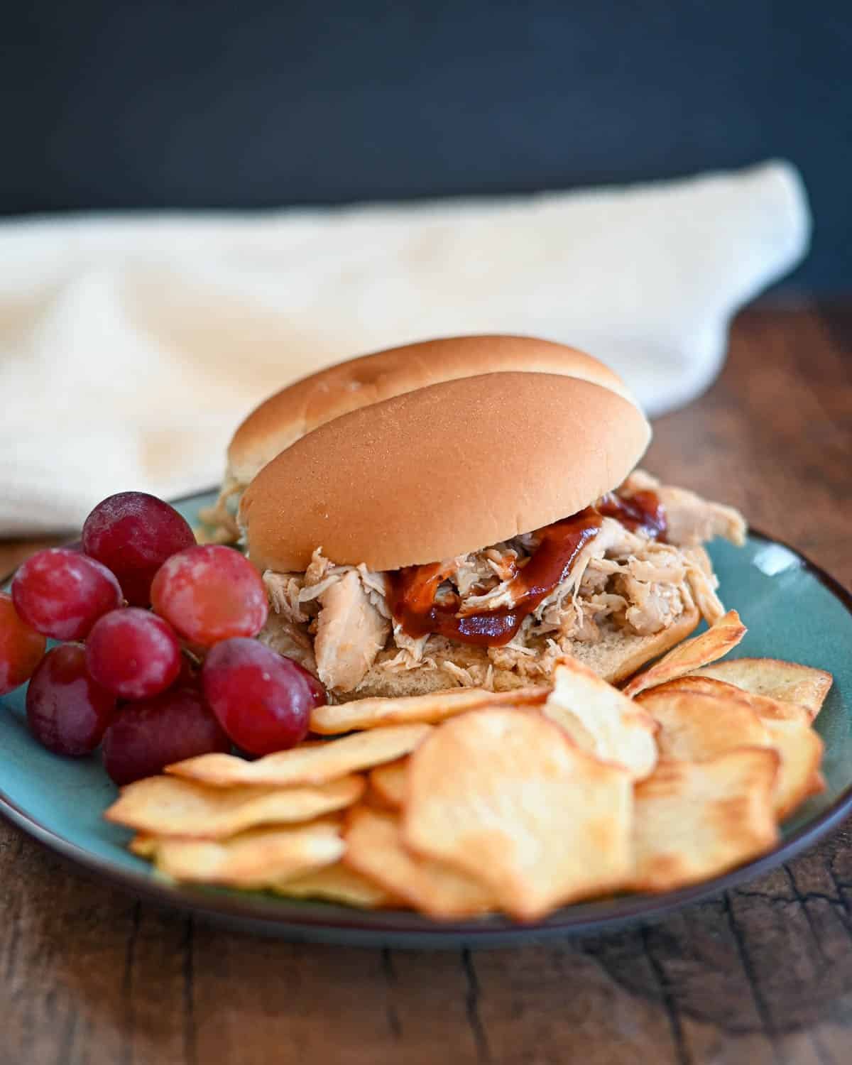 A shredded chicken sandwich next to chips and grapes on a plate.