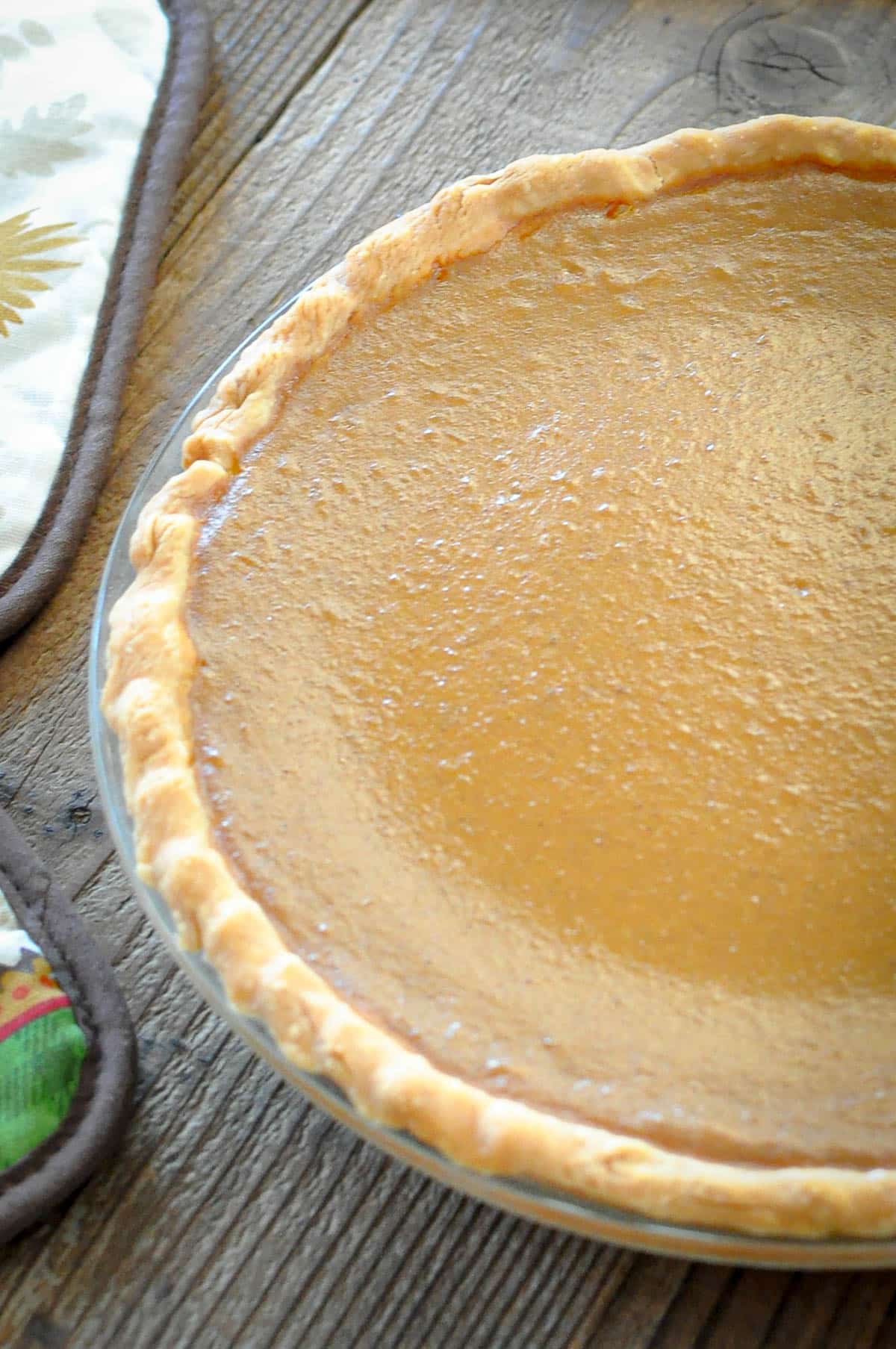 A homemade Butternut Squash Pie in a glass pie dish sits on a wooden table next to oven mitts.