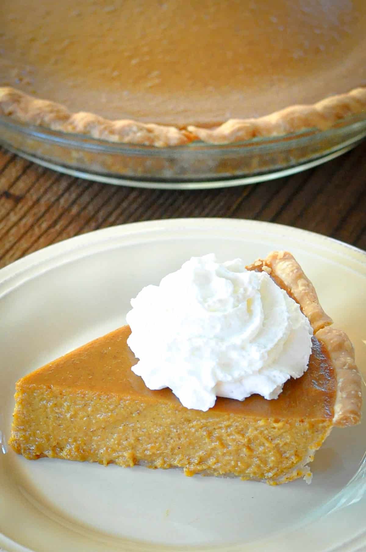A slice of Butternut Squash Pie with whipped cream on top, served on a white plate, with the rest of the pie in the background.