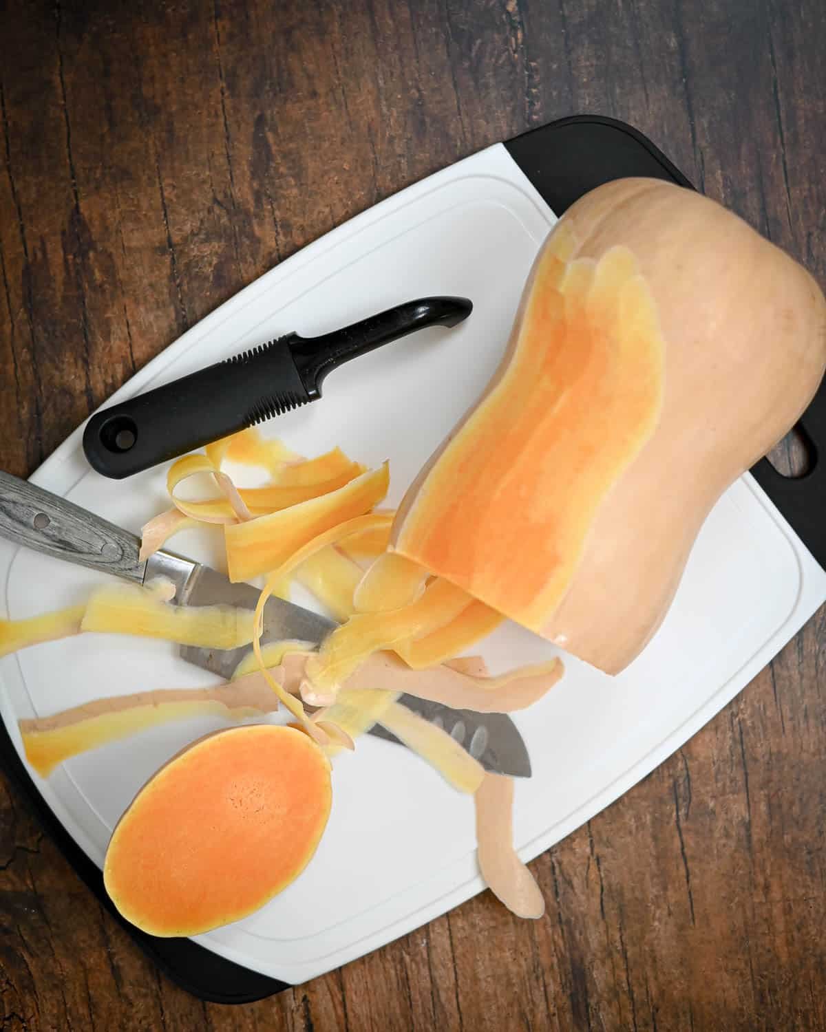 Butternut squash being peeled with a vegetable peeler on a white cutting board, next to a knife-perfect prep for your homemade Butternut Squash Pie.