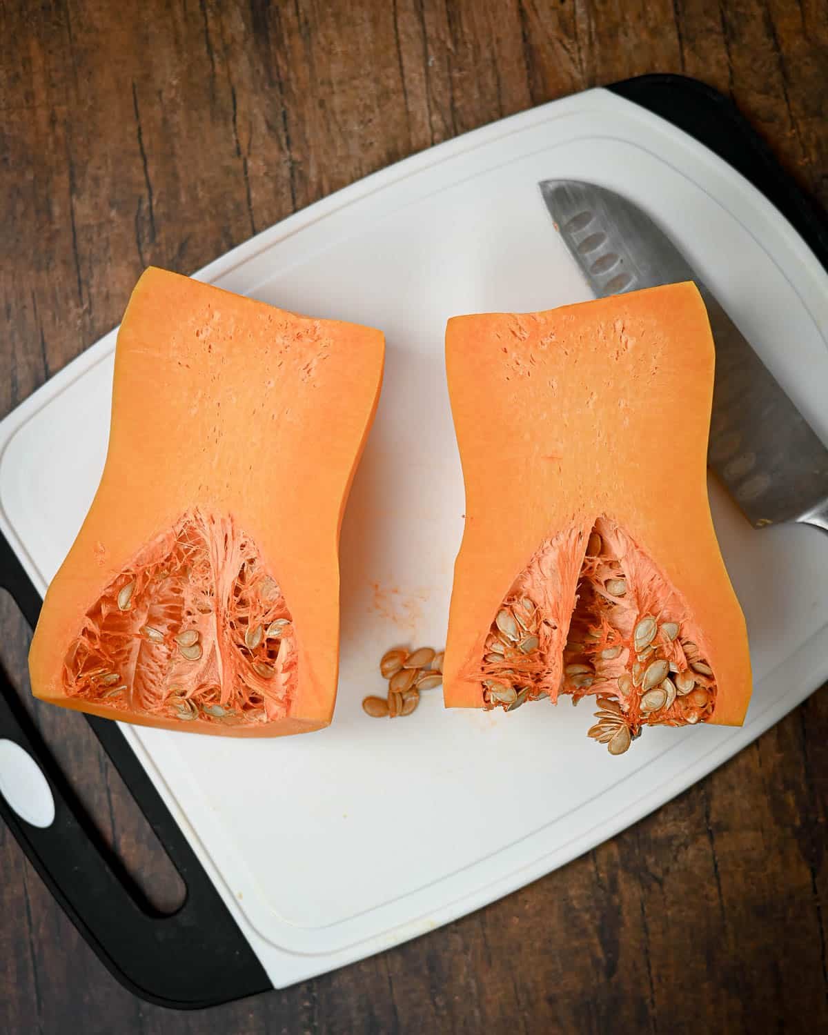 Halved butternut squash with seeds on a white cutting board next to a knife on a wooden surface, perfect for making Butternut Squash Pie.