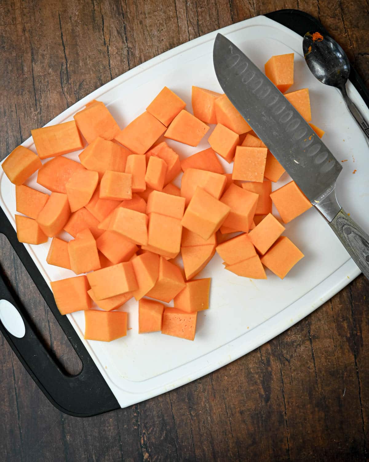 Cubed sweet potatoes on a white cutting board alongside a knife and spoon rest on a wooden surface, ready to be used in a delicious Butternut Squash Pie recipe.