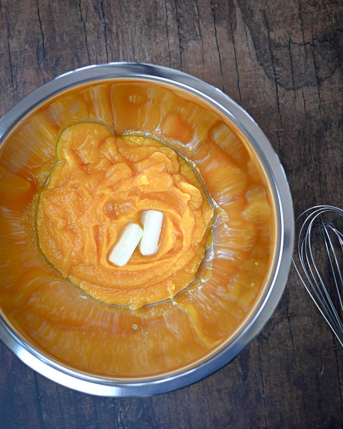 A metal bowl with pumpkin puree and butter, ready for a Butternut Squash Pie, sits next to a wire whisk on a rustic wooden surface.