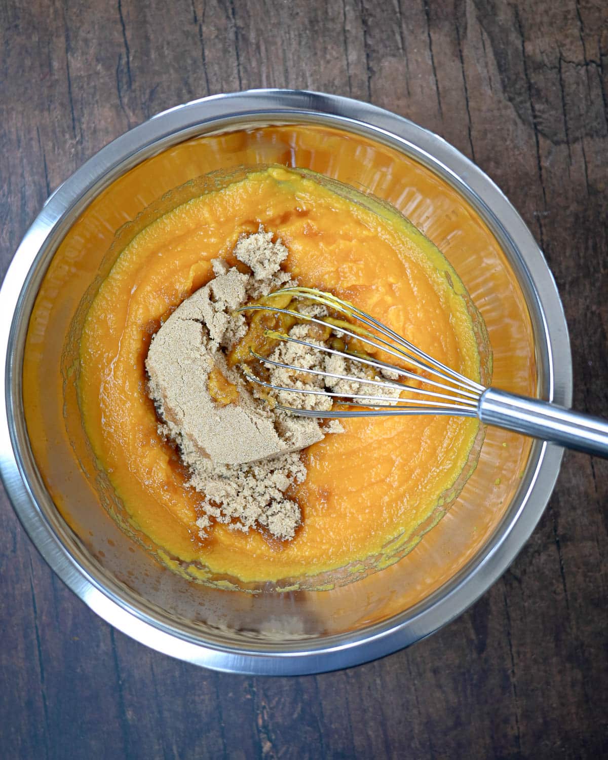 A whisk mixing brown sugar into orange puree in a stainless steel bowl on a wooden surface, prepping the filling for Butternut Squash Pie.