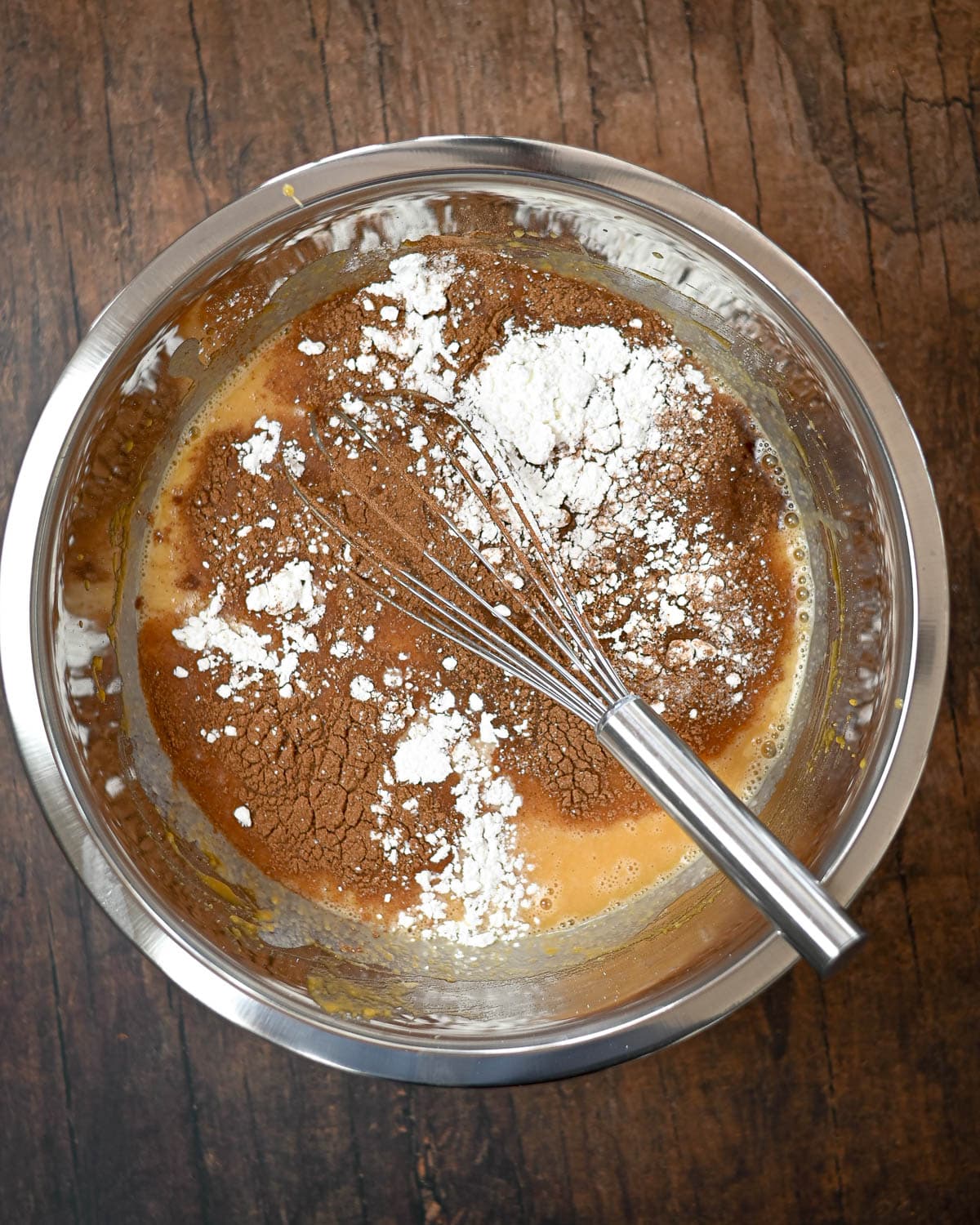 A mixing bowl with wet and dry ingredients for Butternut Squash Pie and a whisk on a wooden surface.
