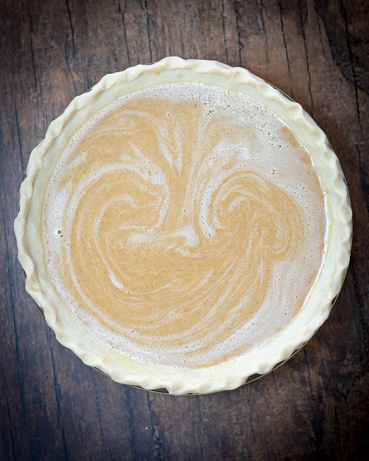 Unbaked Butternut Squash Pie with light brown swirled filling and crimped crust rests on a rustic wooden surface.