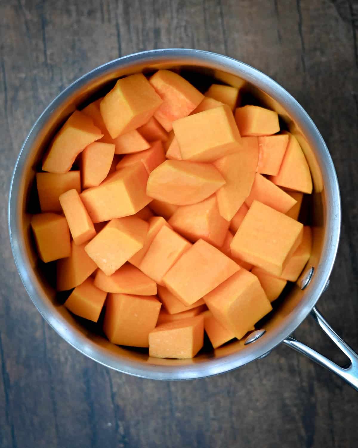 Cubed butternut squash in a stainless steel saucepan on a wooden surface, ready to be used for a delicious Butternut Squash Pie, viewed from above.