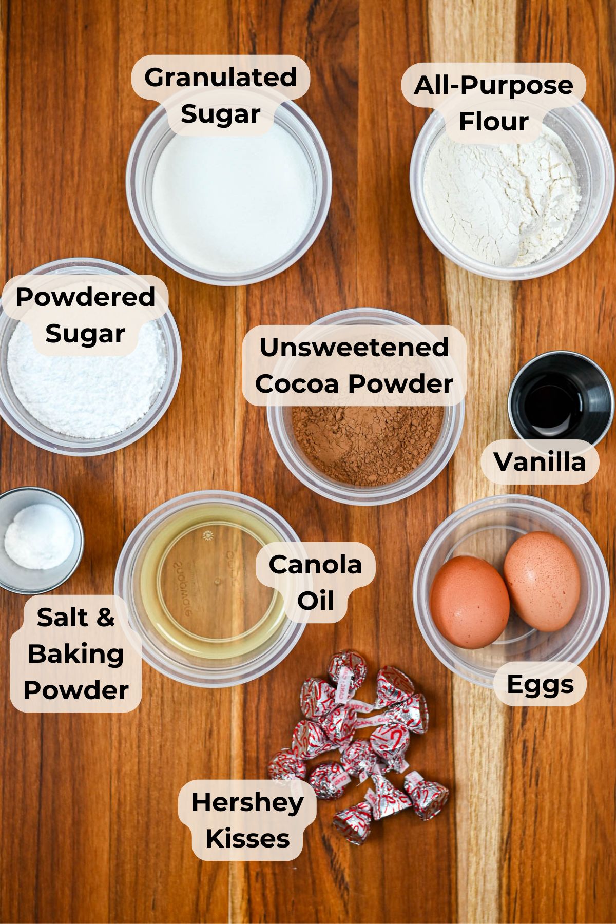 Baking ingredients in bowls, labeled: sugar, flour, cocoa, oil, vanilla, eggs, powdered sugar, Hershey Kisses, salt-everything you need to make delicious Kiss Crinkle Cookies or classic chocolate crinkles.