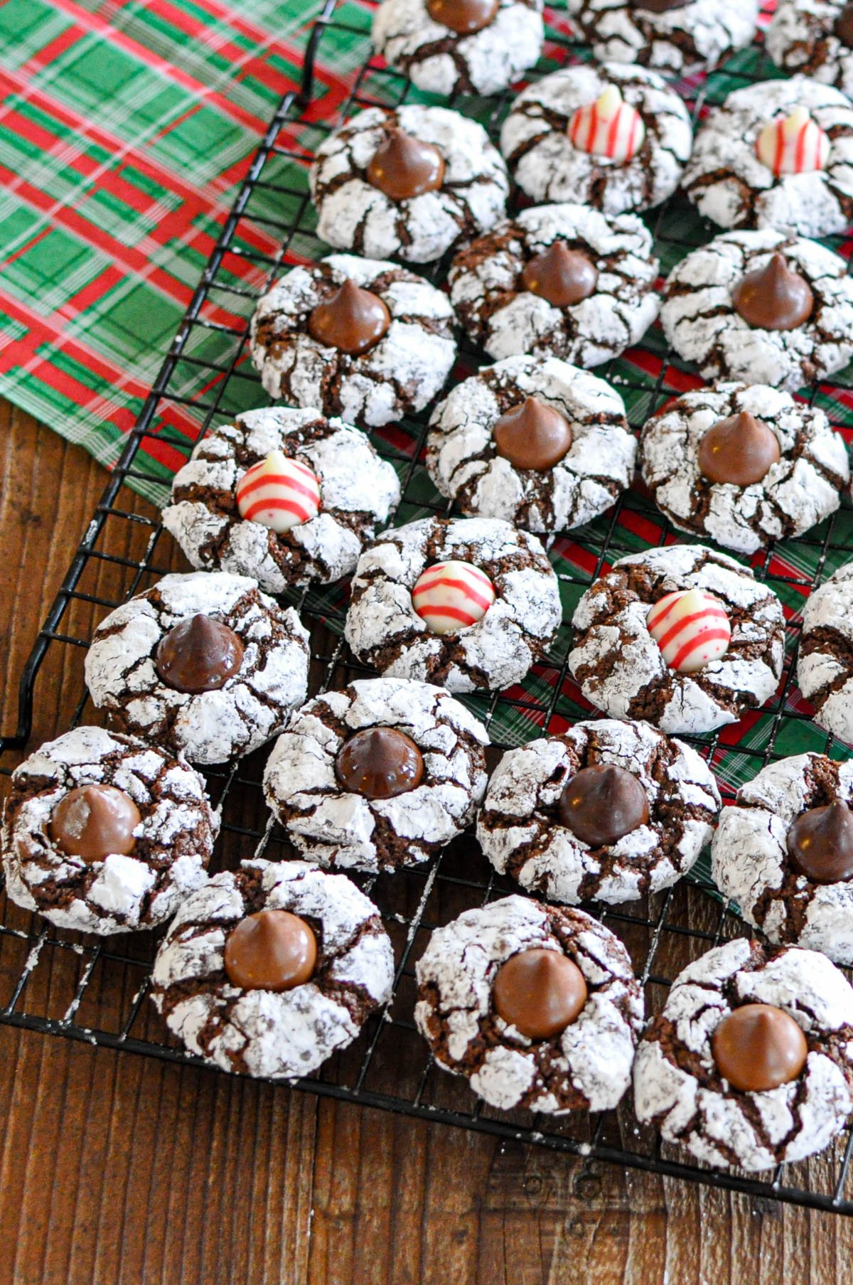 Kiss Crinkle Cookies topped with chocolate and peppermint kisses rest on a cooling rack, with a plaid cloth nearby, showcasing the classic charm of chocolate crinkles.